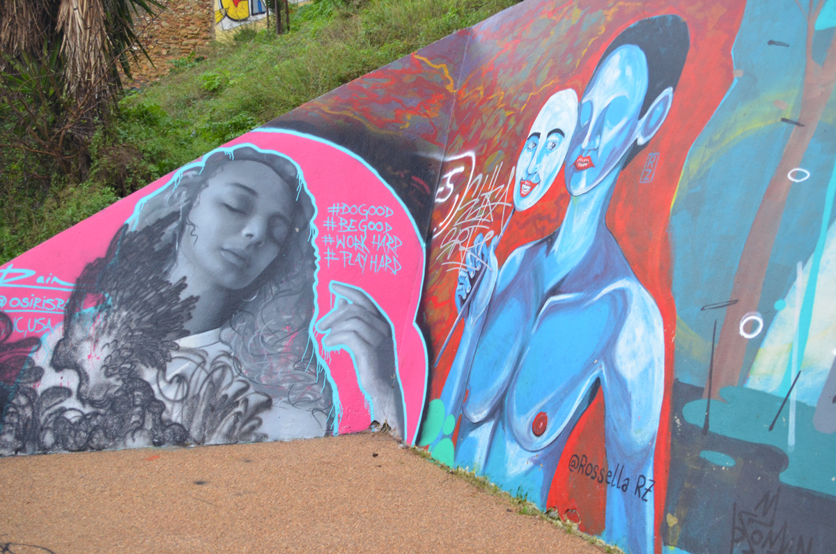 two murals on a wall, on the left in shades of grey a woman with long curly hair and eyes closed with words #begood #doggod #playhard #workhard.  on right is blue person with blank face holding a mask, also in blue, with a happy face