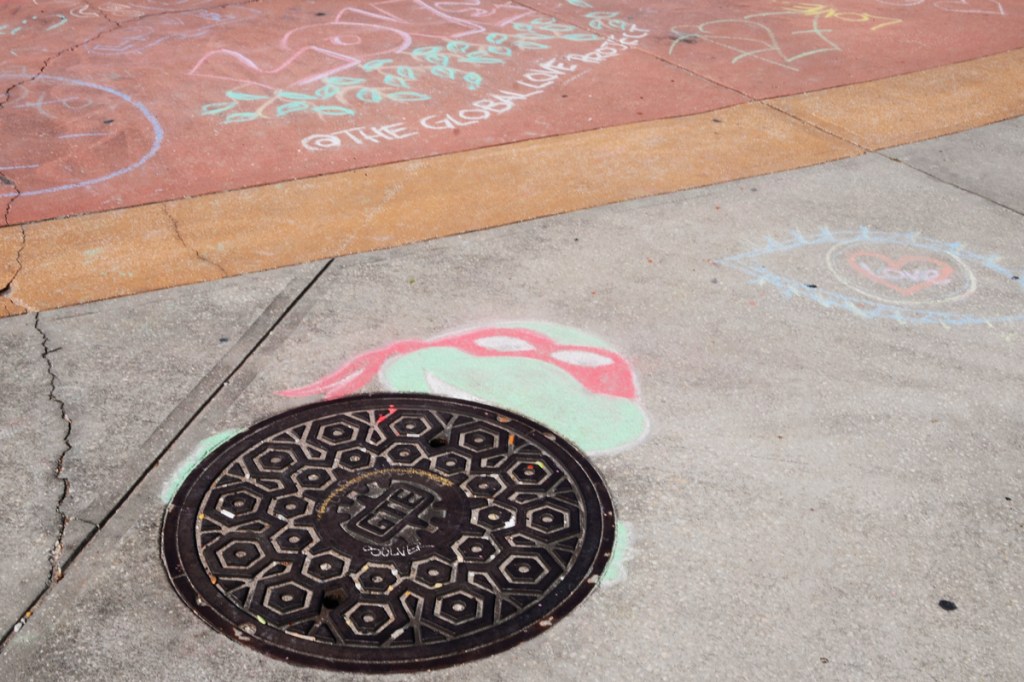manhole cover on the sidewalk, chalk drawing of head and shoulders of teenage mutant ninja turtle with a red mask, Raphael