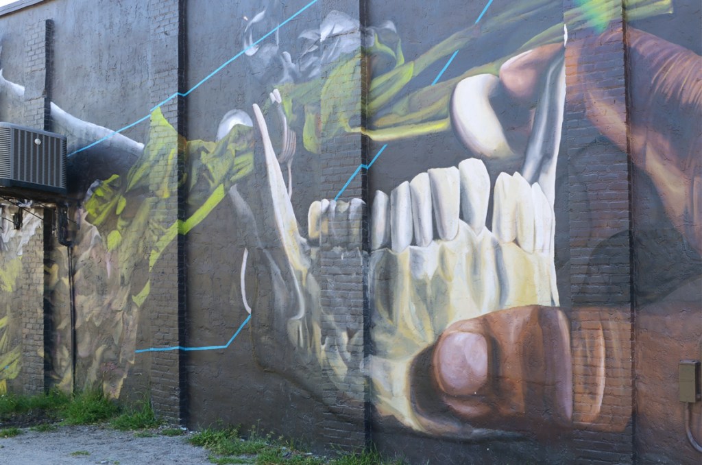 mural by Bakpak Durden a large jaw from a skull with white teeth, a hand is reaching out to touch it 