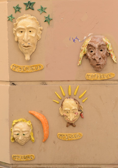 4 faces, 3 D, made of plaster, painted, on an outside wall, street art, labelled as Mr. Serieux, Mr. Affreux, Mme Lune and Mr. Soleil.