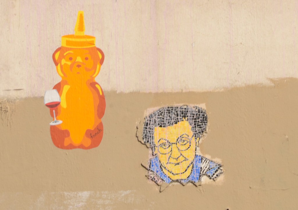 two graffiti pieces on a wall, a honey bear with a glass of red wine and a tile portrait