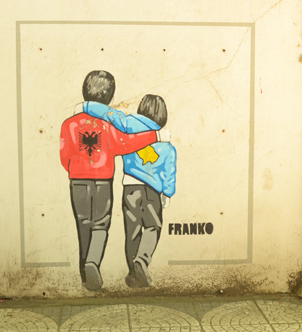 mural painted by franco on a wall in Tirana, of two young boys, backs to painter, one wearing a red jacket with Albanian symbol, the other wearing a blue jacket with yellow Ukranian map on it 