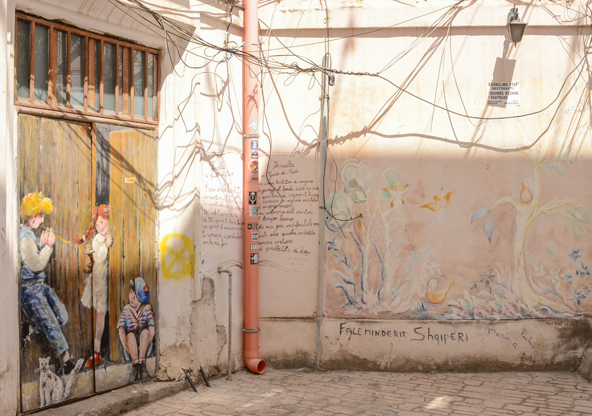 paintings in a laneway, on the left is a painting of a partially open wood doorway with a girl stepping out and smiling at the two boys on the front step. A black and white cat is also there. Some words are written in the corner, then on the right is a pale pastel coloured painting of flowers and leaves