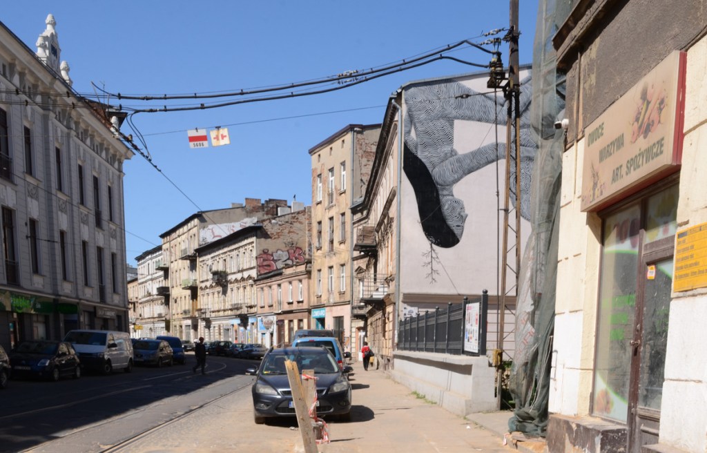 street scene in Lodz Poland with parked cars, sidewalk, stores, and other buildings, a mural is partially visible