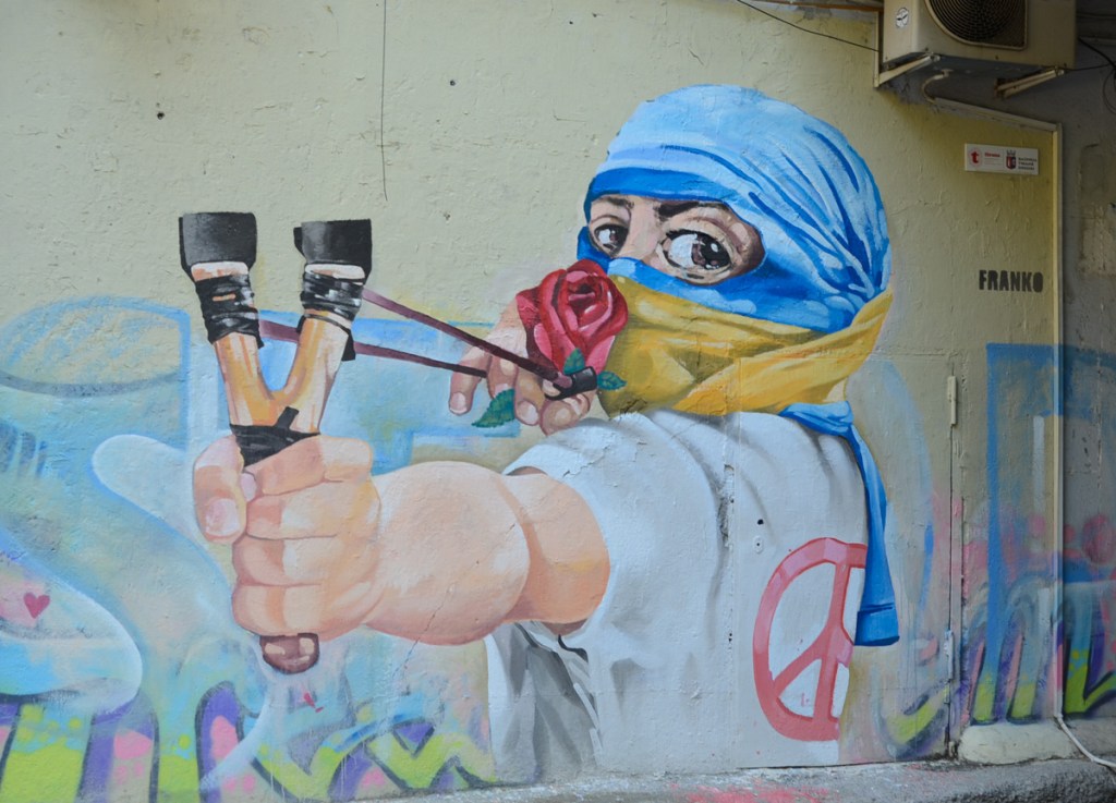 mural of a young person with mask partially covering the face mask is made using Ukranian flag, shooting a sling shot