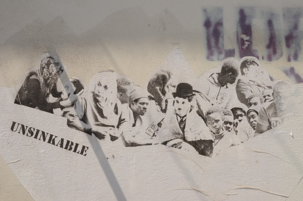 black stencil on white wall with heads of many famous people, Laurel and Hardy, plus word unsinkable