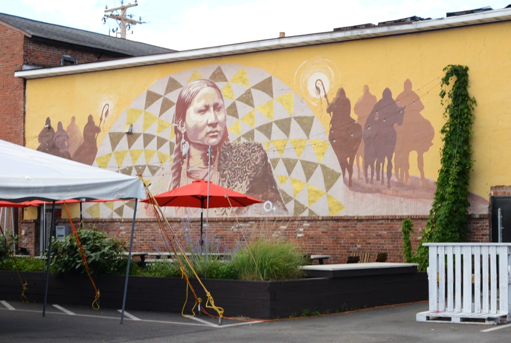 mural, center is first nations portrait surrounded by circles of triangles looking like the rays of the sun, silhouettes of people on horseback on either side