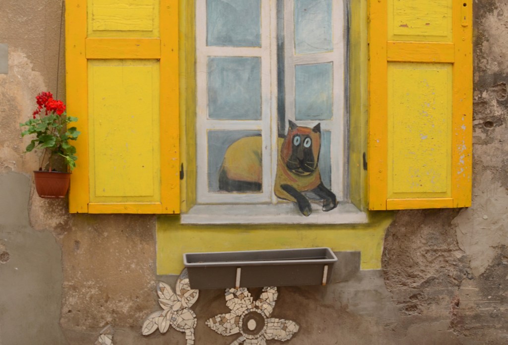 bright yellow shutters on a window, pot of geraniums, red geraniums, beside, an empty flower box below, a painting of a fat brown cat half out the window and half inside