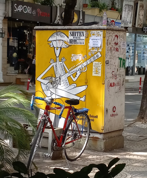 painted sidewalk box on a sidewalk in Tirana, Sargent Pepper cartoon character in yellow playing guitar while sitting cross legged on the floor