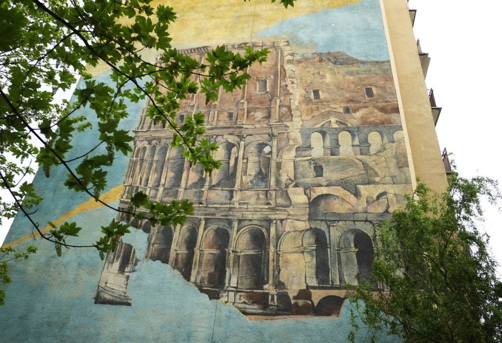 with some large trees nearby, a large mural of the ruins of the Roman coliseum in Rome, or any other roman coliseum for that matter