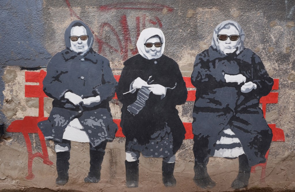 street art on a concrete wall, three older women sitting on a bright red bench. Women are wearing grey winter coats, head scarves, and sun glasses. the woman in the middle is knitting