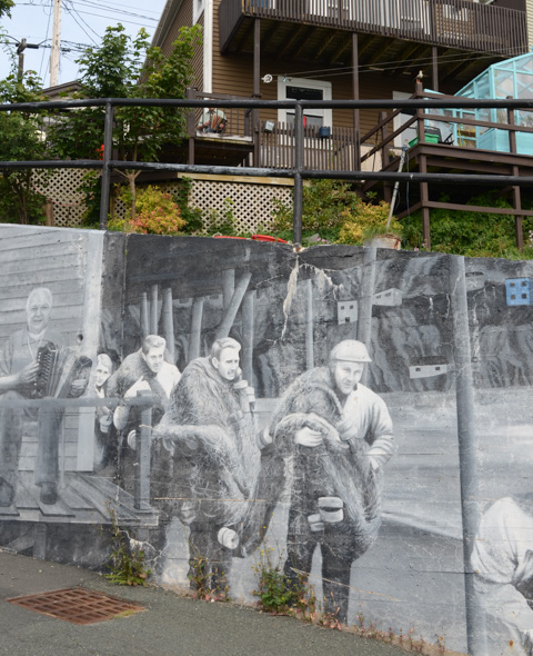 the back of brown wood house with a retaining wall, concrete, at the bottom, painted with mural with scenes of historic Battery neighbourhood fishing community