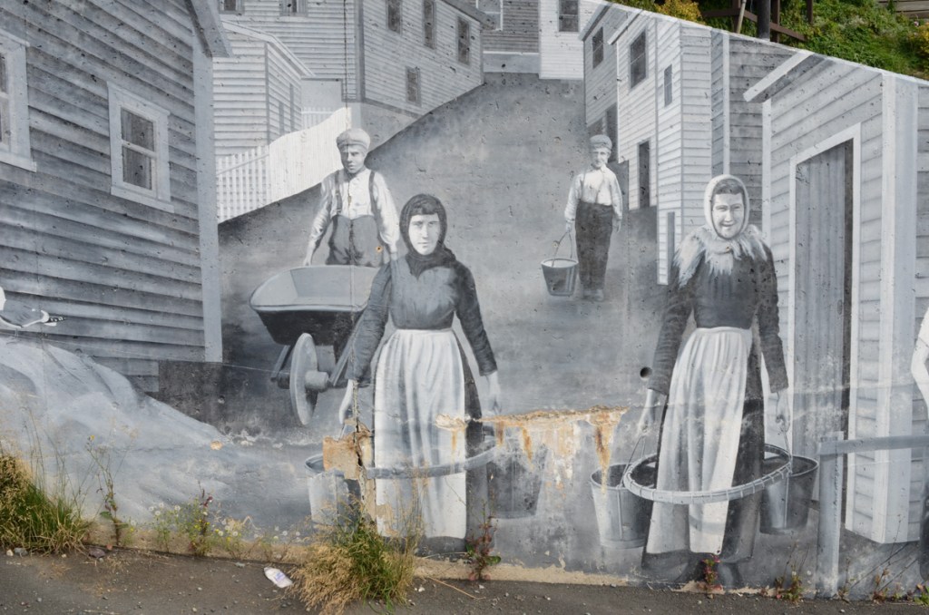 part of a mural in grey tones, black and white, showing narrow street with two people carrying buckets or pails and another person with a wheelbarrow