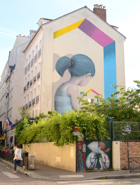 large mural painted by seth on the end of a row of buildings on rue Mouffetard in Paris, a girl with a ponytail is drawing a simple box-like building with different coloured crayons. 
