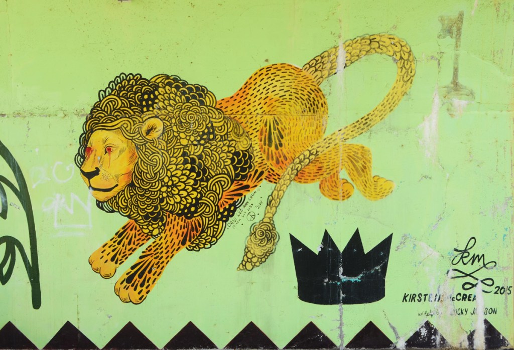 a street art painting of a lion with a curly mane and long tail, painted by Kirsten McCrea in yellow and black