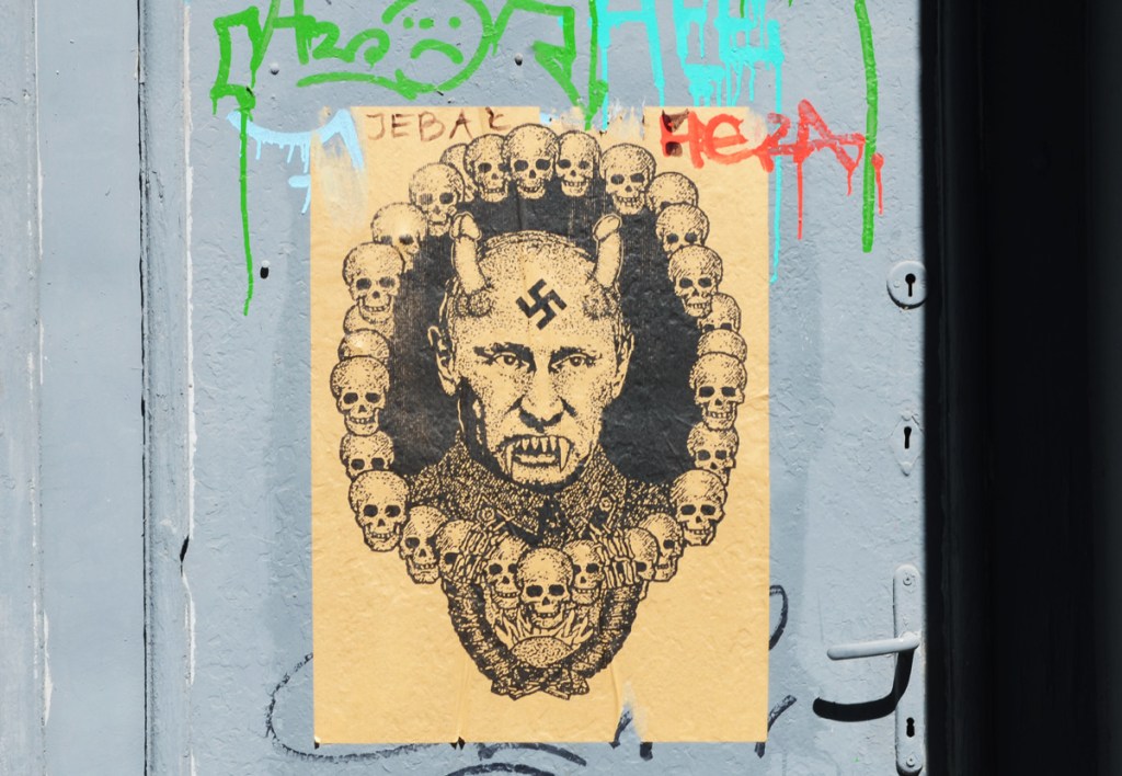 paper pasteup graffiti with image of Putin with Nazi swastika on his forehead, fangs for teeth, and phallic devil's horns on his head.  He is surrounded by a ring of small skulls