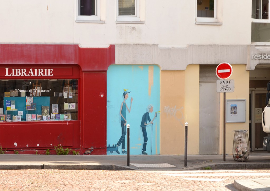 red storefront, librairie (book store) with a small mural painted on the wall beside it, a painting by Oji of an older grey haired person with a cane spray painting something on a wall, and a younger man with a yellow bird on his head and pulling a wagon, watching.