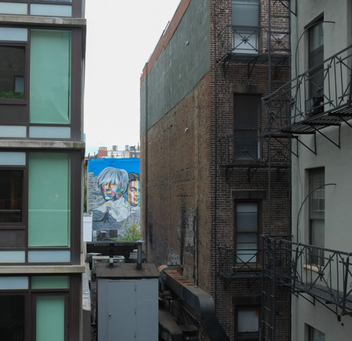 glimpse of a mural with portrait of Andy Warhol in the background, seen in a gap between two taller brick buildings