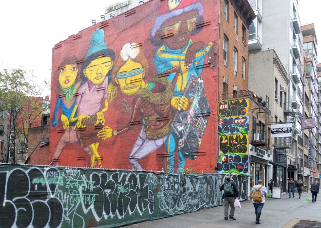 two men walk past a vacant lot with a tagged fence in front of it, on the far side of the lot is a large Osgemeos mural hip hop homage on 14th street in New York city, young people with ghetto blasters playing music and dancing 