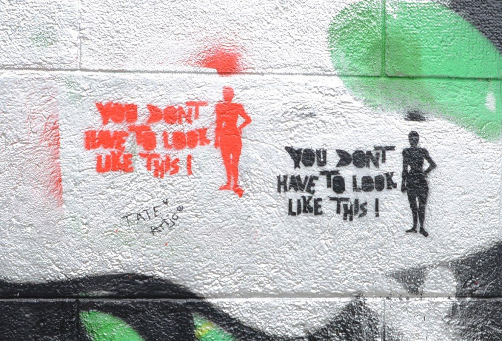 two stencils on a concrete wall, one red and one black, both with a silhouette of a person's figure and both with words that say you don't have to look like this 