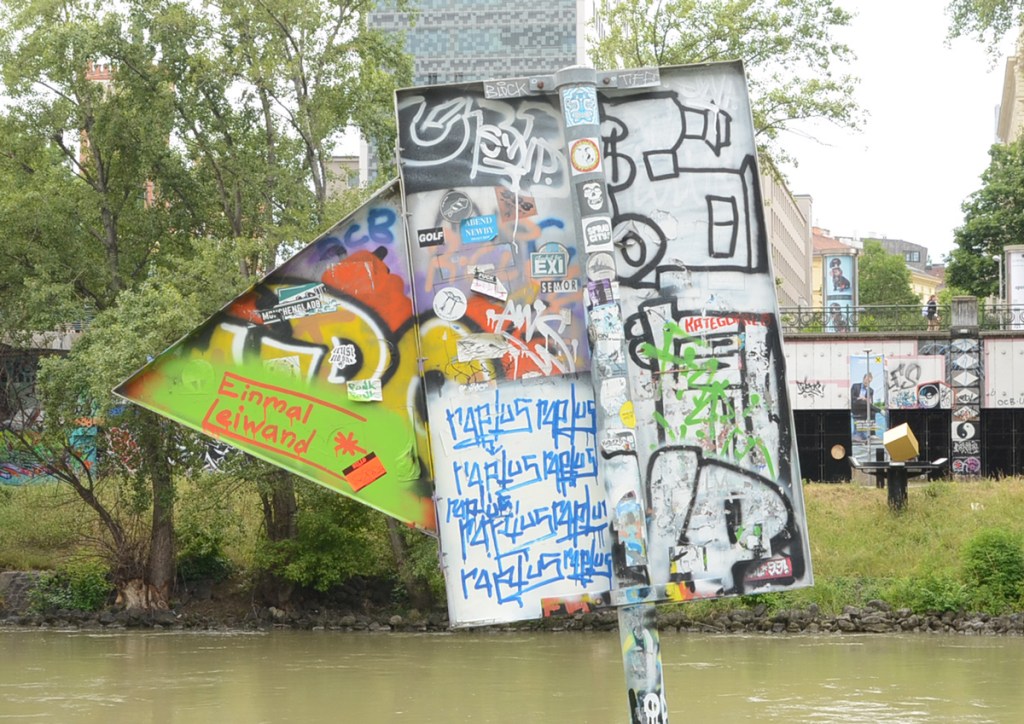 directional sign by the waterfront that has been covered with stickers and some painted graffiti tags 