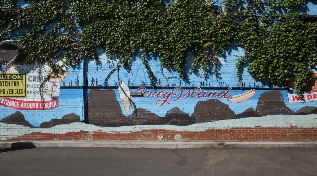 blue mural with coney island in cursive, an ince cream cone and a hot dog, but ivy is growing from above and starting to cover up the mural