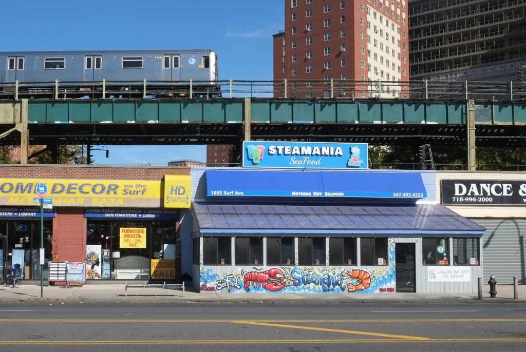 subway is above ground, and above the stores along a coney island street, near Steamania Seafood restaurant and its shrimp mural along the sidewalk