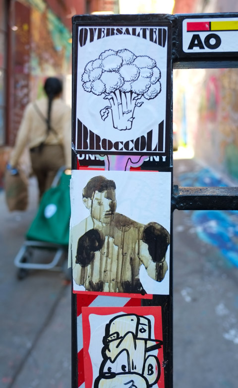 a woman pulling a green bag walks past a pole with three stickers on it. on the top is unsalted broccoli a bunch of broccoli, in the middle is an image of a shirtless boxer wearing boxing gloves