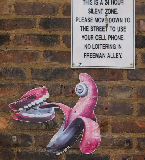 wheatpaste on a brick wall, a pink and purple partially peeled banana with one eye on the tip has part of its peel inside an open set of dentures (or an open mouth) also in pink and purple, by eye sticker graffiti artist. Above it is a sign saying no noise and no loitering in the alley, Freeman Alley