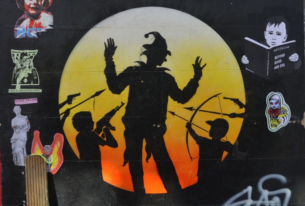 Graffiti on a wall, wheatpaste graffiti surrounds a large orange and yellow circle with black silhouettes inside, boyes with rifles, spears, and bows and arrows pointed at man with jester hat and arms raised 