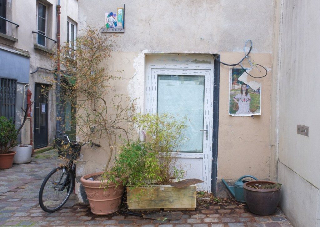 in a Paris alley, a white backdoor with plant pots beside, a poster on the wall on one side and a small graffiti high on the other side