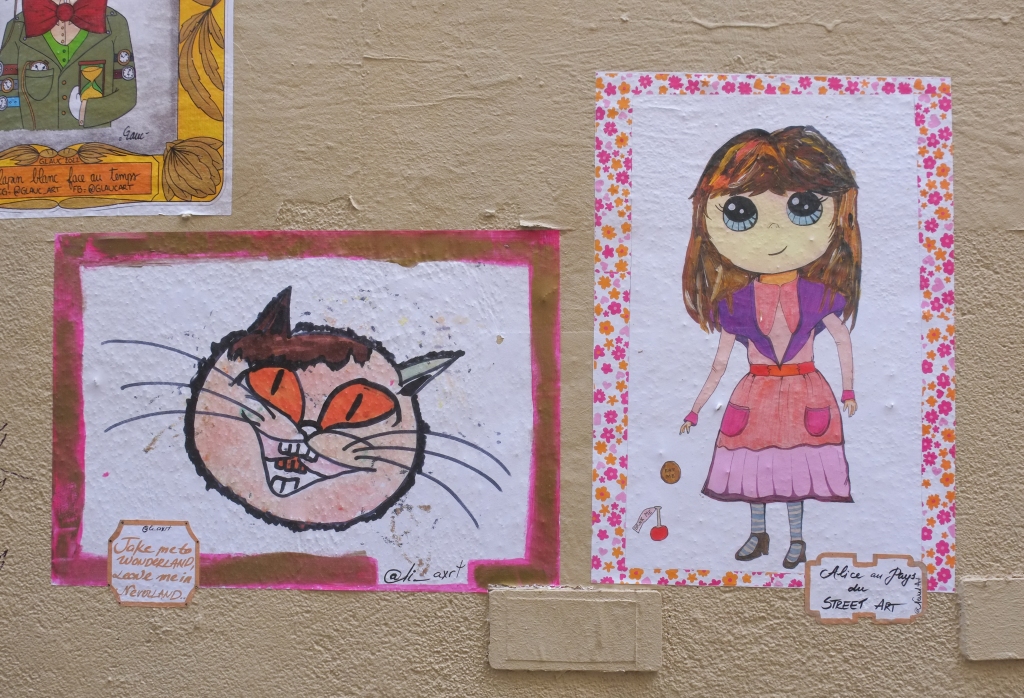 2 poster on a wall, one is a large picture of a cat face and the other is a girl in pink and purple, Alice