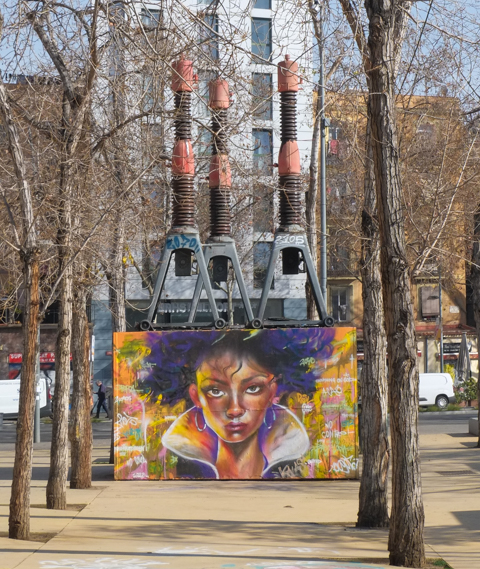 in a park, trees on both sides, a large wall painted with a mural of a womans face, urban part, old pieces of equipment from a power plant act as sculptures behind the wall