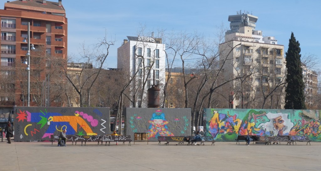 urban park in Barcelona, tall buildings in the background along with leafless trees (early spring), concrete space with large murals on one side, people sitting on benches in front of murals