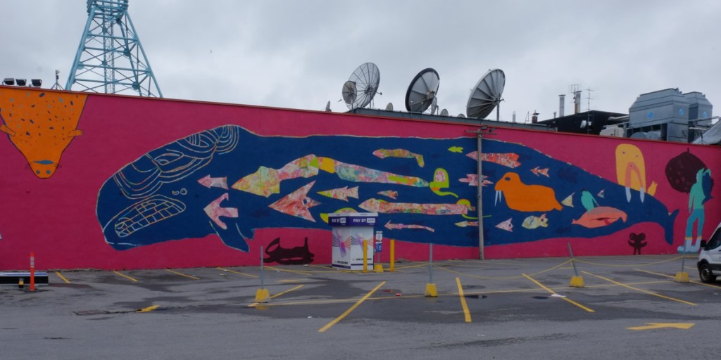 large horizontal mural on a wall beside a parking lot featuring a blue whale on a pink background.  Some other marine animals are also in the mural