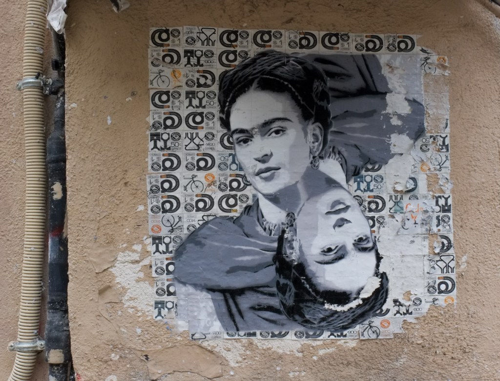 paper wheatpaste graffiti on a wall in Paris, looks like picture is printed on background of Mexican export stamps, or Mexican postage stamps. Frieda Kahlo
s portrait, 2 copies, radially symmetrical, in grey tones, smiling, hair tied back