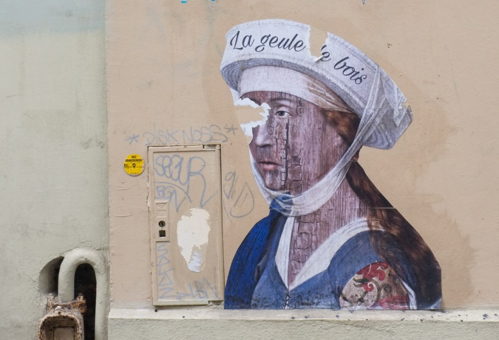 wheatpaste street art on a wall, a person with long dark hair, blue sleevelss shirt, a tattoo on shoulder, white head band and white hat strapped to head. the hat has words la geule de bois