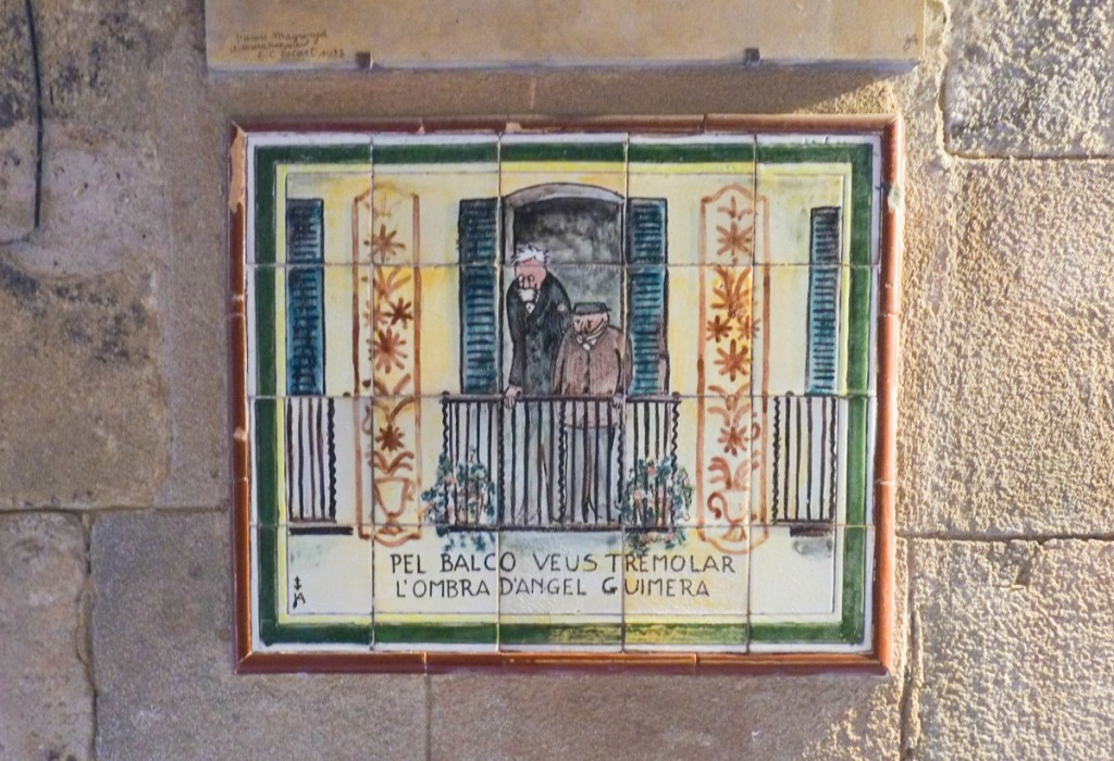 small ceramic picture on a wall, with catalan text Pel balco veus tremolar l'ombra d'Angel Guimera, picture is of two men standing on a balcony, blue shutters beside window, 