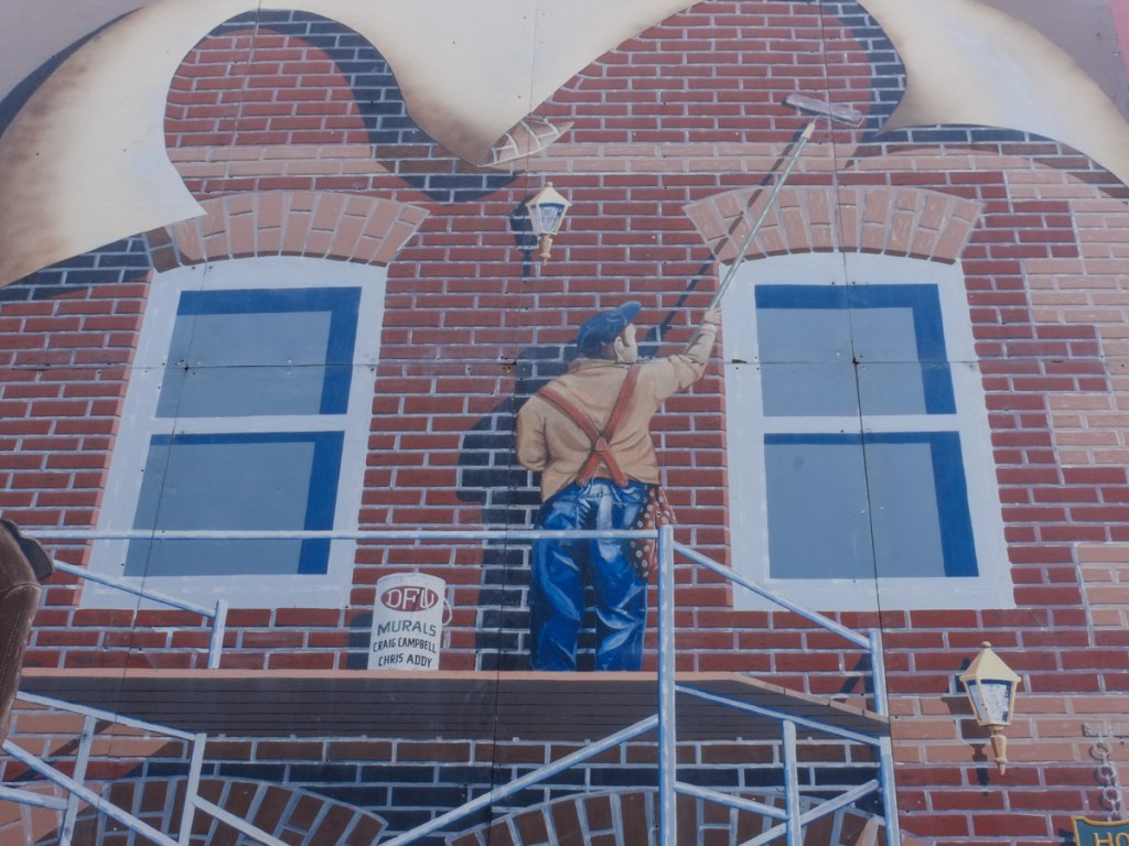 mural showing man painting the front of a two storey brick office building, standing on scaffolding
