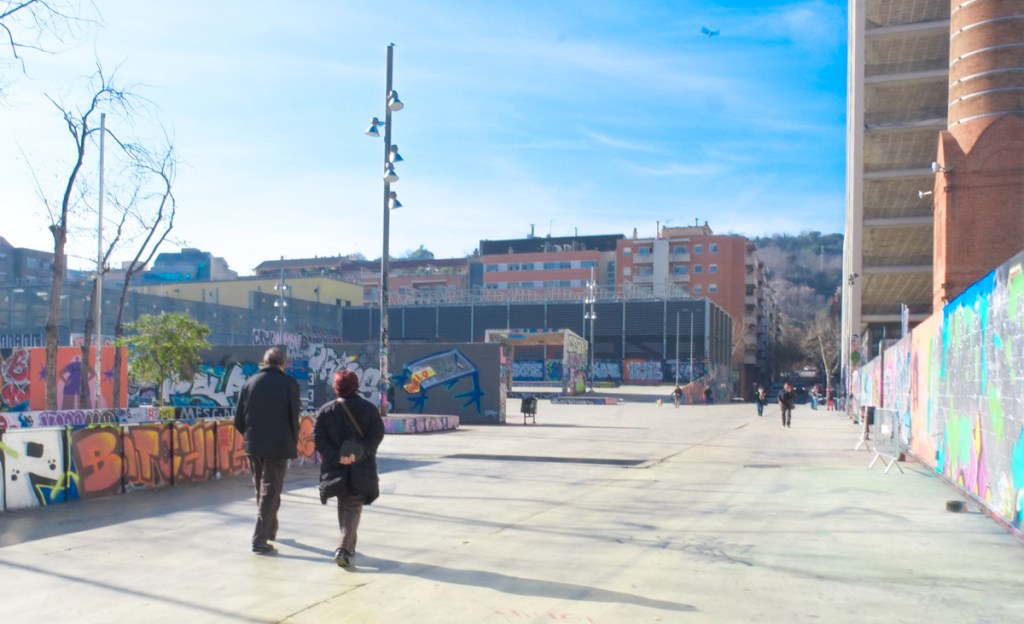 two people walking through an urban park, on a concrete path, walls of street art around them, residential buildings of Barcelona in the background