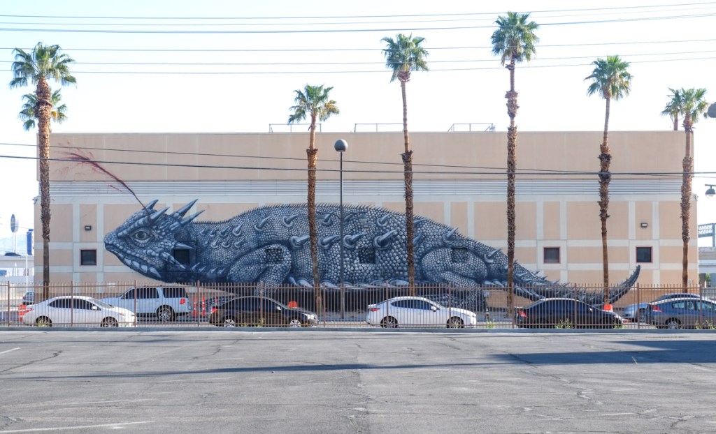 giant treet art mural on the side of a building, in grey tones, a giant horned lizard, with 6 skinny palm trees in front as well as some cars parked in front
