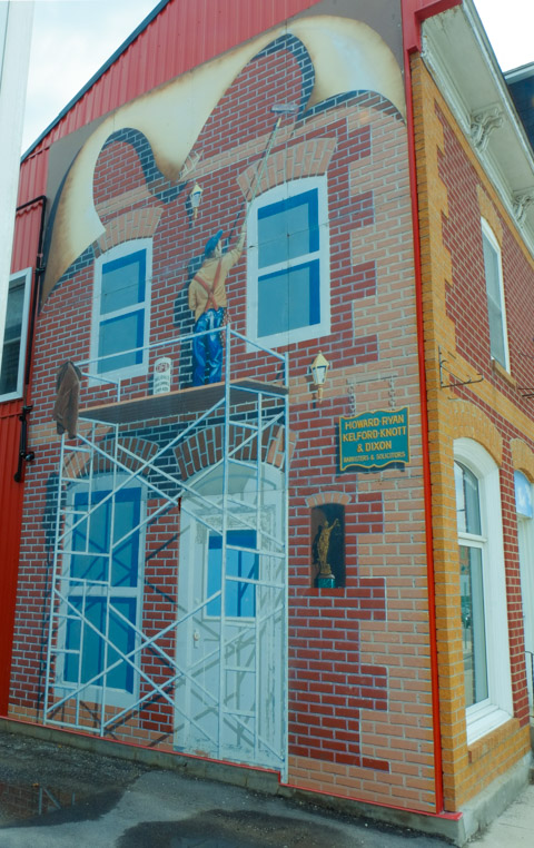 mural showing man painting the front of a two storey brick office building, standing on scaffolding