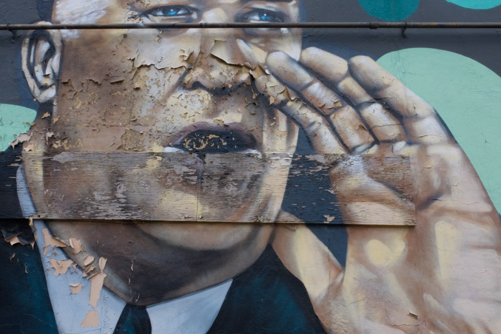 close up view of mural, man with hands by mouth as he is calling, paint peeling on the concrete and wood