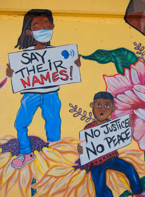 close up of part of mural, a girl holding a sign that says say their names and a boy with a placard that says no justice no peace. 