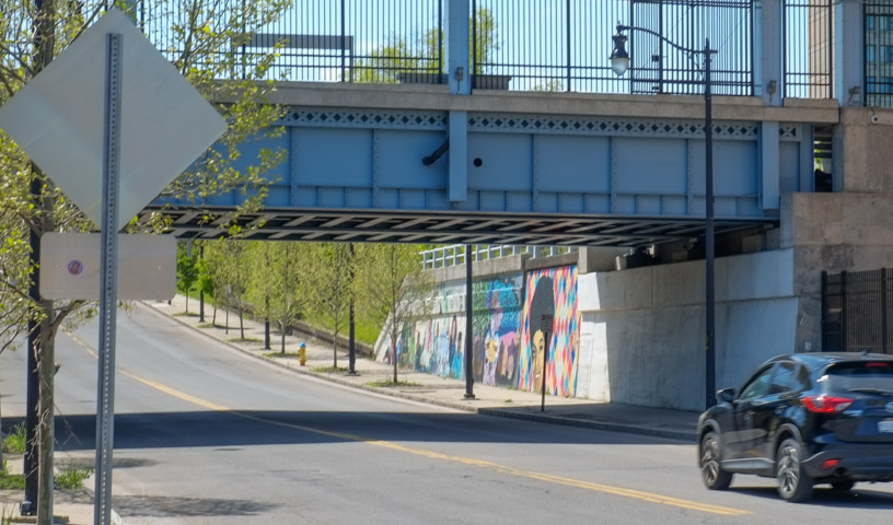 a smal black car drives under a railway bridge beside amtrak station, murals on the side of the underpass