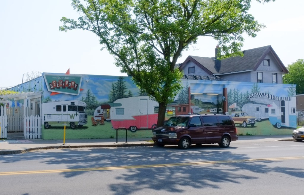 a van is parked in front of Rochester Beer Park with its's campground mural on the wall, WInnebago, vintage campers and trailers are parked there, motorcycles, old station wagon, camp chairs set up by a table