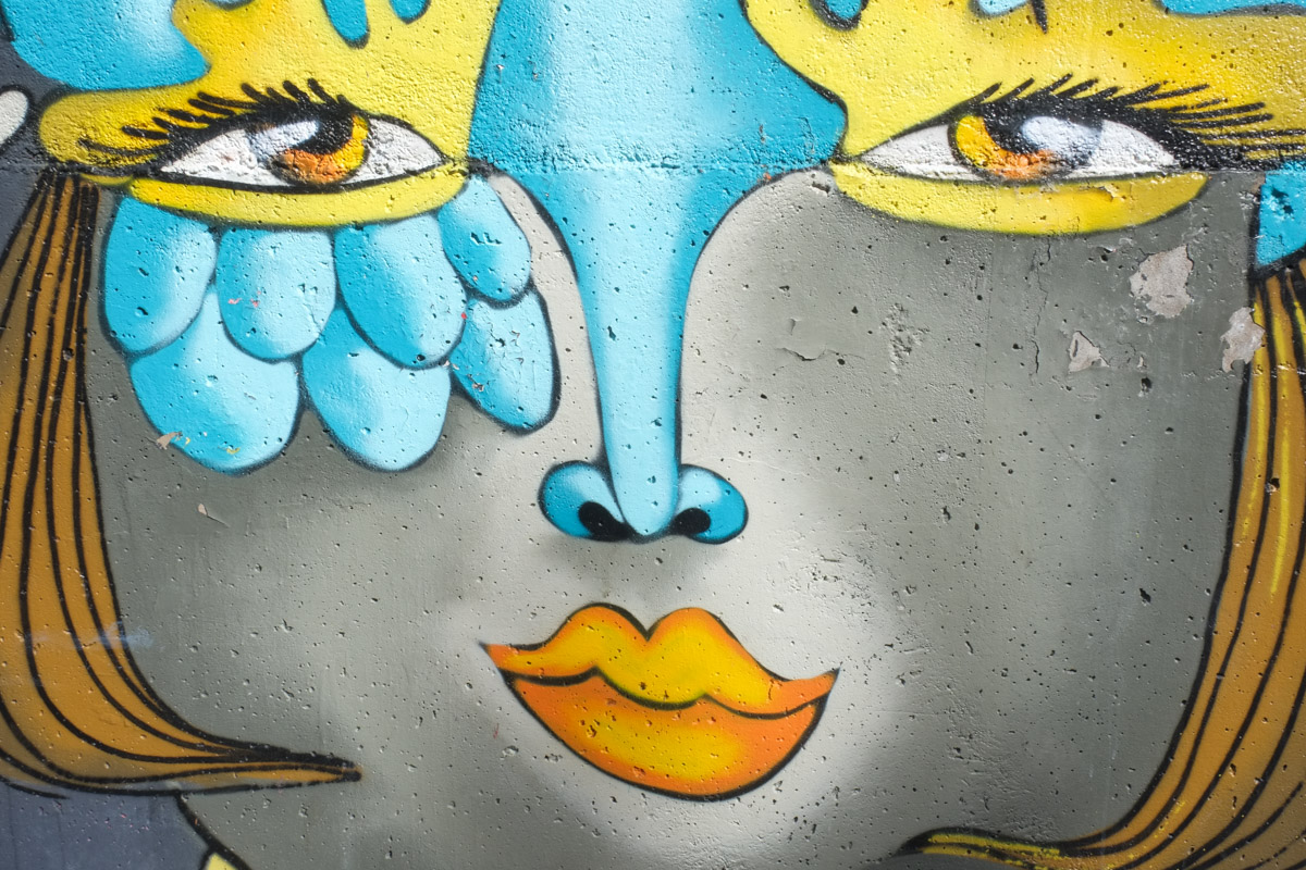 close up of a face in un kolor distinto mural, on grey concrete, a face with blue nose, gold hair, and orange lips