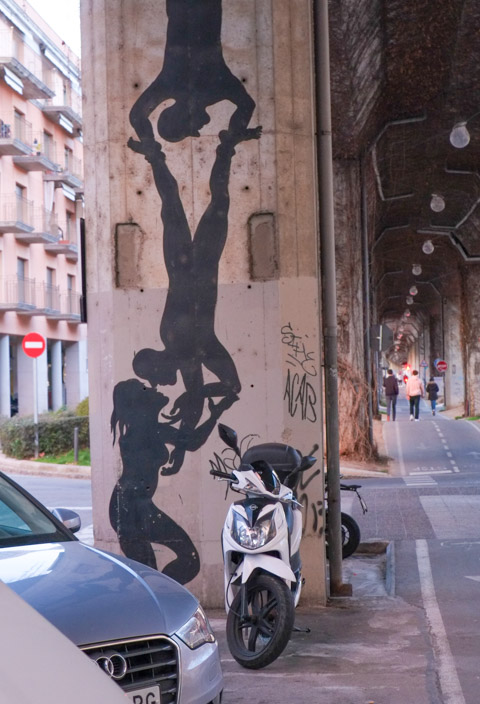 silhouette black people in street art, on a concrete pillar, top is man hanging upside down holding the feet of another man who is also upside down and kissing woman standing on the street, motorcycle parked in front