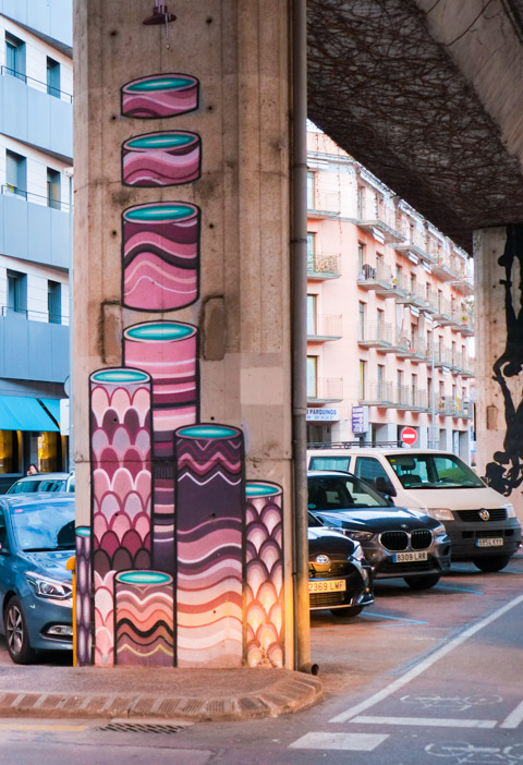 street art painting by kram on a concrete post, cars parked behind, painting is sliced stacks of patterns