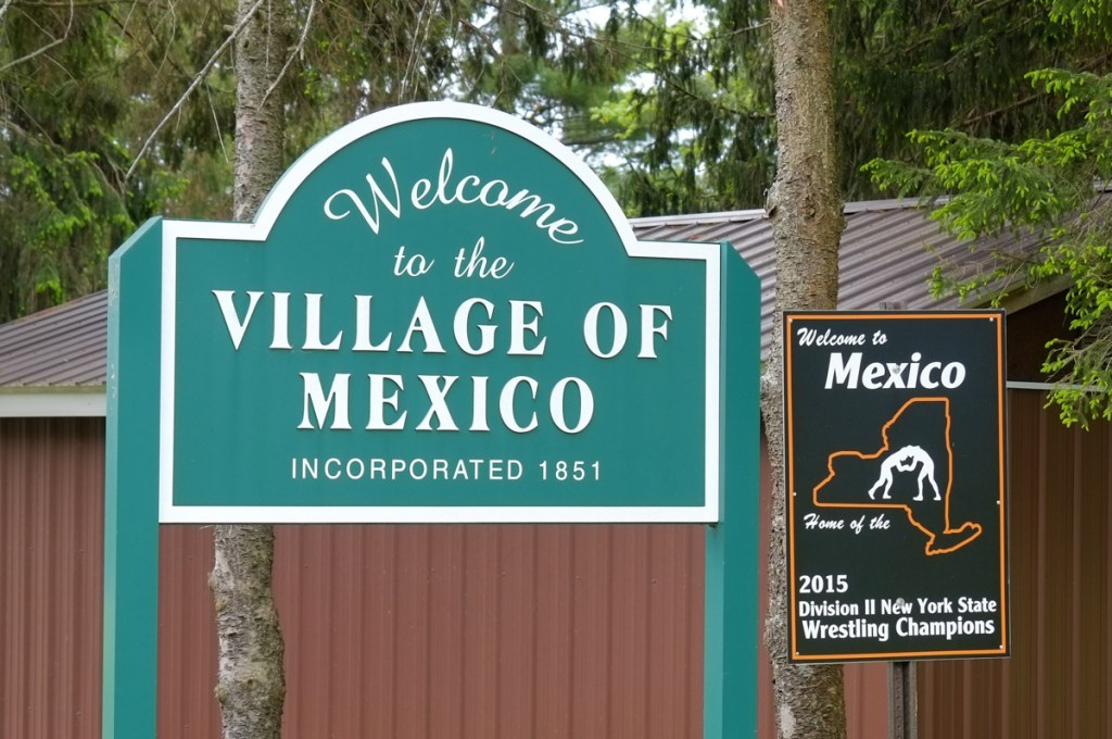 large green and white sign that says welcome to the village of mexico, a smaller sign beside it that claims